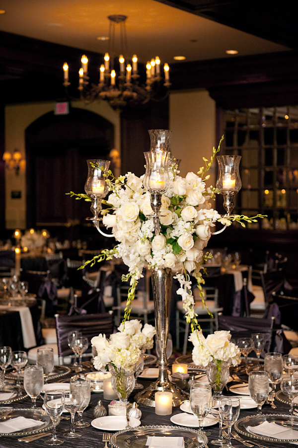 Beautiful reception table setting - large silver candelabra with ivory candles and white and green floral arrangement as centerpiece, silver dishes, and dark purple tablecloth and chair covers - photo by Houston based wedding photographer Adam Nyholt
